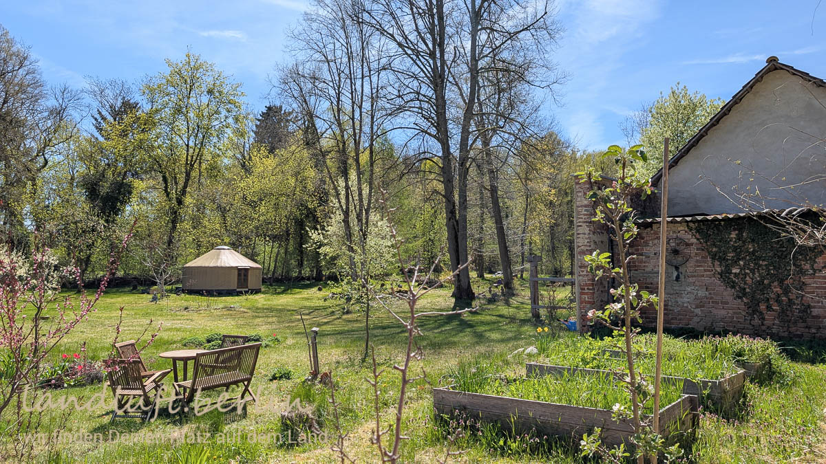 Blick aus der geschützten Ecke in den Garten, vorne die Hochbete, hinten die Jurte, rechts Garage und Scheune